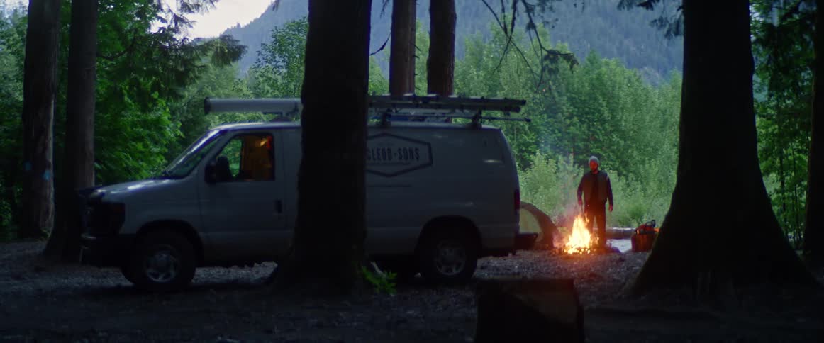 A man pokes a campfire set up next to a commercial van