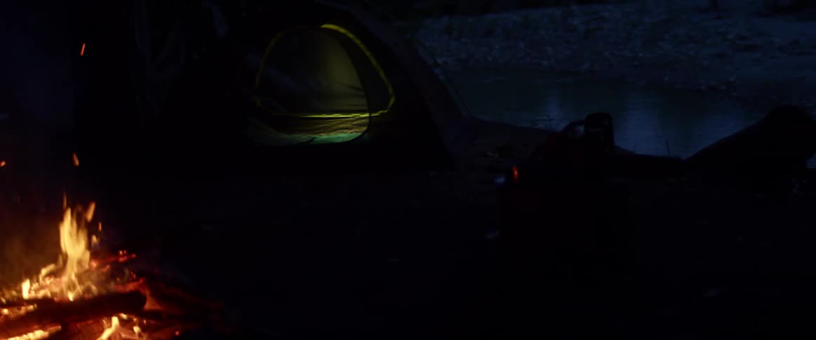 A campfire burns outside of a tent in wilderness at night
