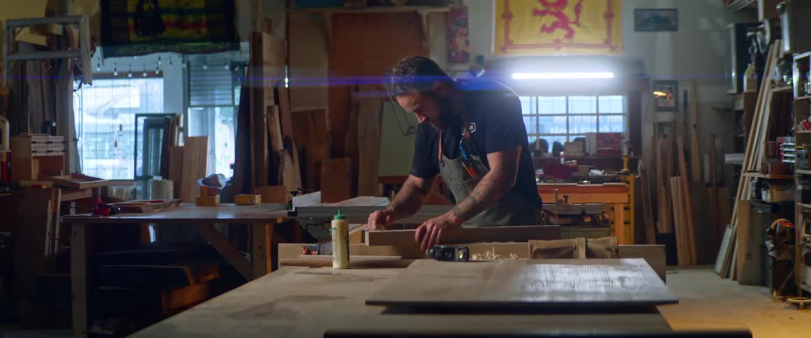 A man in a workshop with an apron working on a piece of wood on a table