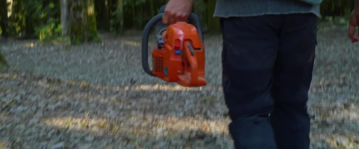 Man with a beanie and a vest carries a saw in the forest