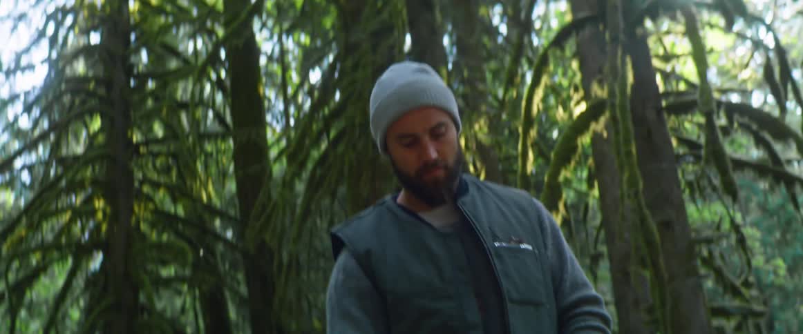 A man repeatedly pulls the rip cord on a chainsaw in an attempt to start it while standing in a forest during the day