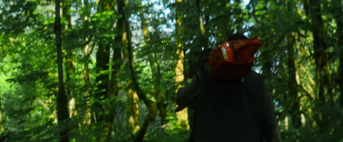 A man walks through a forest while carrying a chainsaw over one shoulder