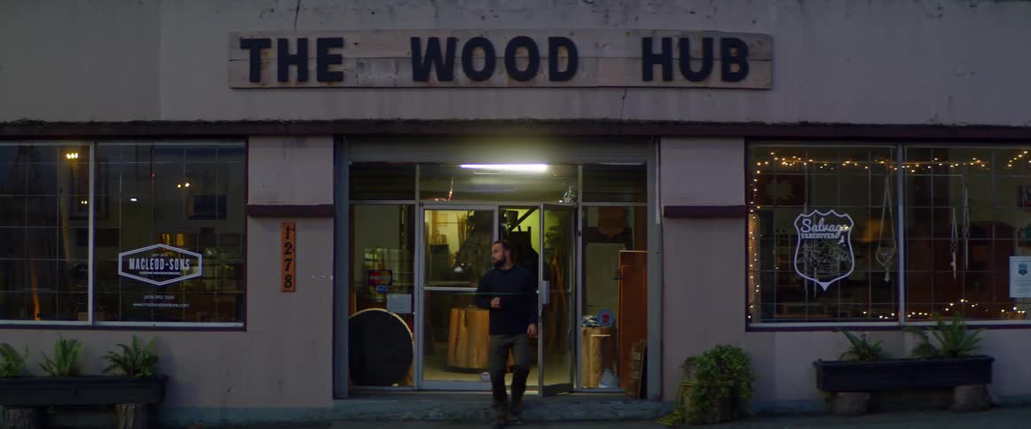 A man walks out the front door of a store that sells wood and looks around outside