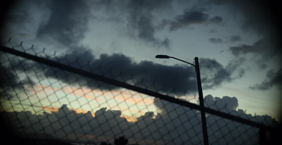 A dark street light on the other side of a chain link fence on a cloudy evening
