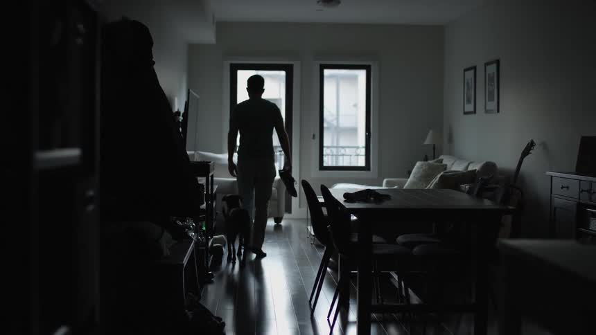 Man walks to living room followed by his dog and sits to put on his shoes