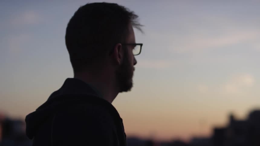 Man with glasses and a beard wandering slowly is silhouetted against the sky at dusk