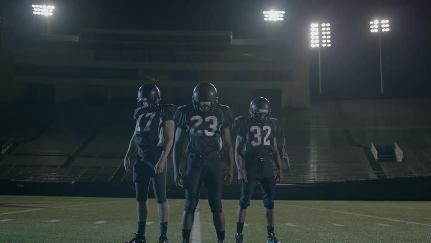 Three uniformed teenagers stand on a football field at night