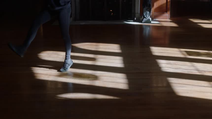 A young woman dances in front of a fireplace as sunlight shines into the otherwise dark room