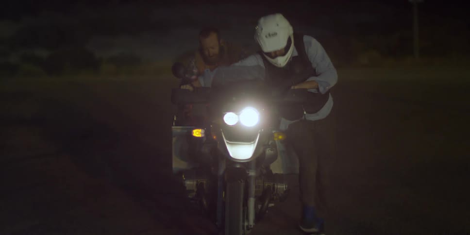 Two men are pushing a motorcycle down a road at night