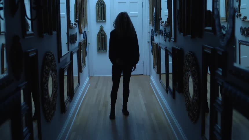 A woman standing and walking backwards in a hallway with mirrors with a door in the background