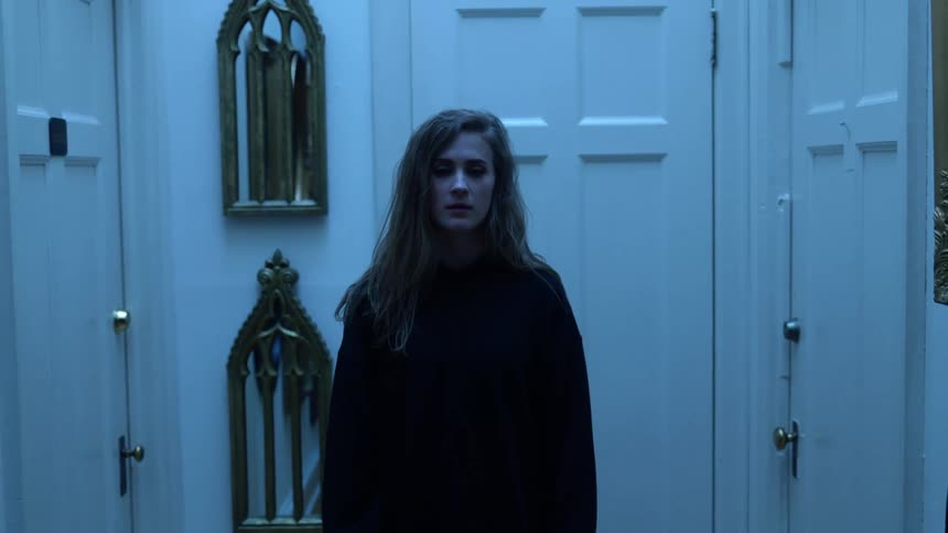 A teenage girl stands in a hallway filled with mirrors with her eyes closed