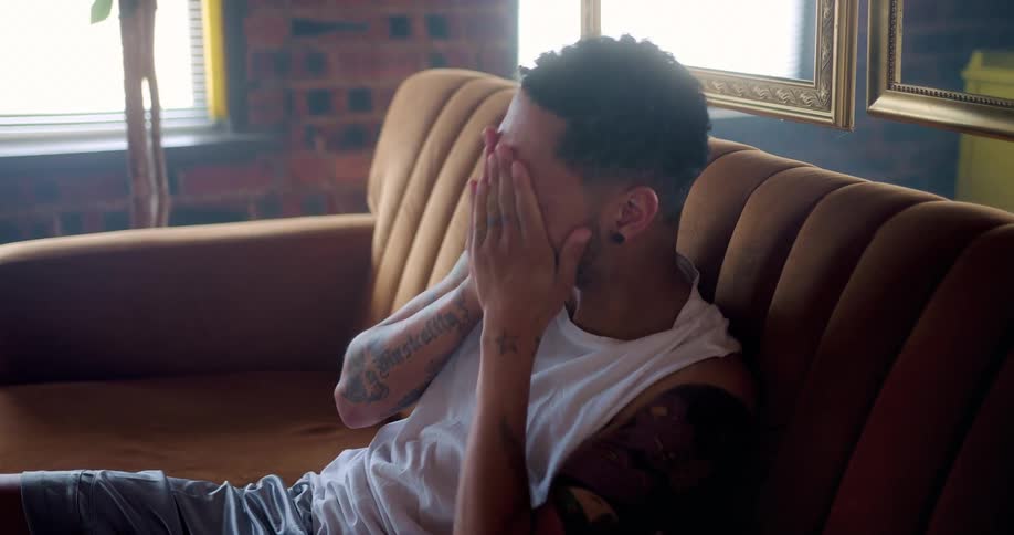 A tired young man rubs his face while sitting on a sofa in a living room during the day