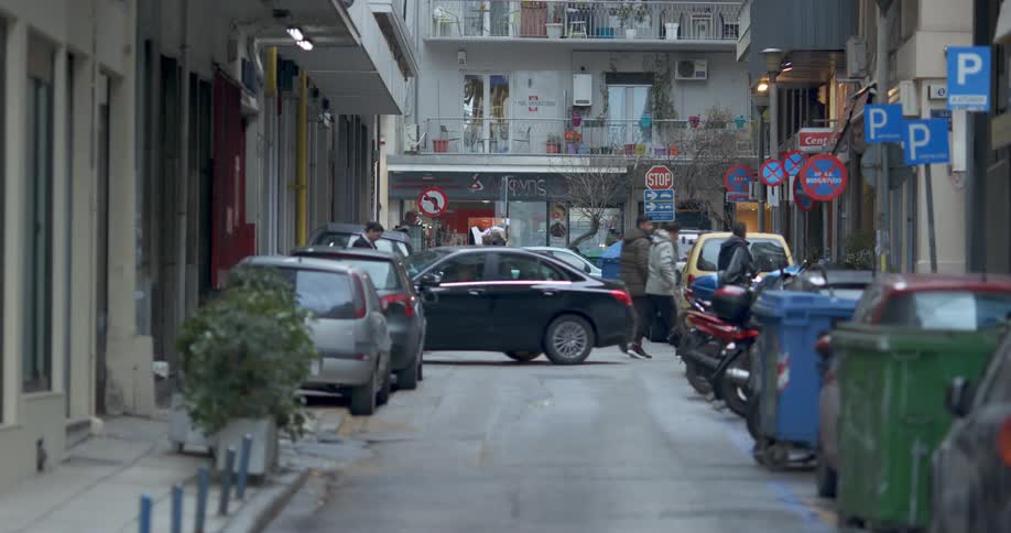 People cross a narrow city street as cars drive by