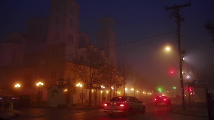 Traffic moving along an intersection next to a large building on a foggy night