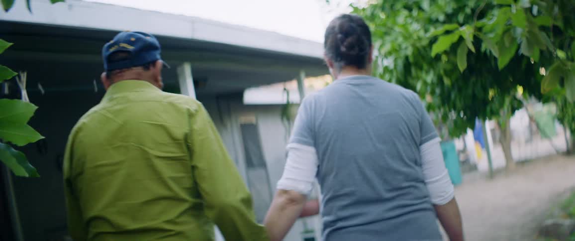 An elderly couple holds hands as they walk along the outside of a small house during the day