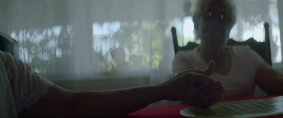 An elderly woman sits at a dining table and another person reaches for her hand
