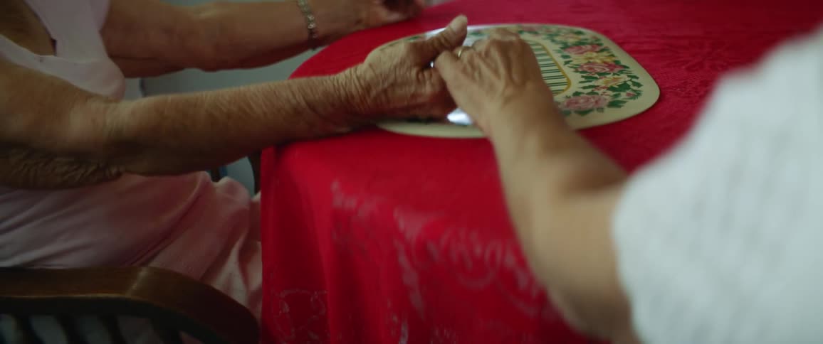 Two elderly women holds hand to pray as they sit down for a meal at a table with a bright red tablecloth