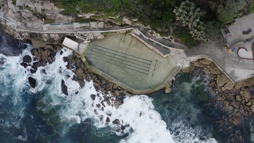 Waves crash against rocks on shore near empty swimming pool and concrete walkways
