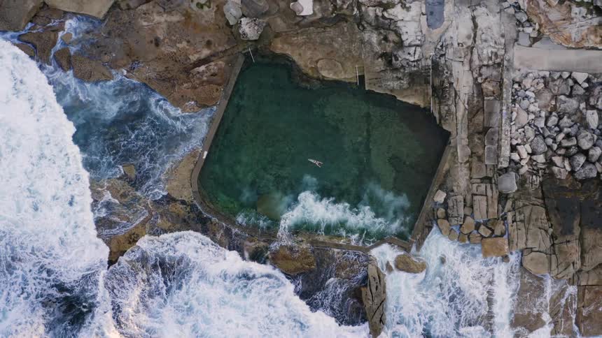 Birds eye view of waves crashing on to the shore with a swimming pool as a person swims in the pool