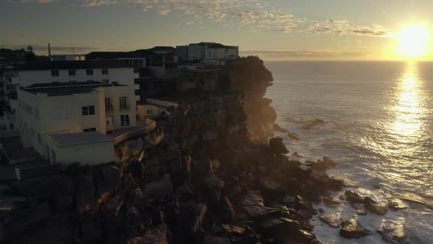 Aerial view of the sun setting over the water as waves roll in to the shore with houses above on a cliff