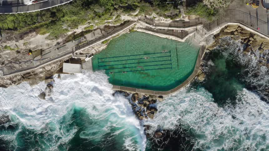 People walk near the shore and swimmers swim through the pool as waves crash against shore