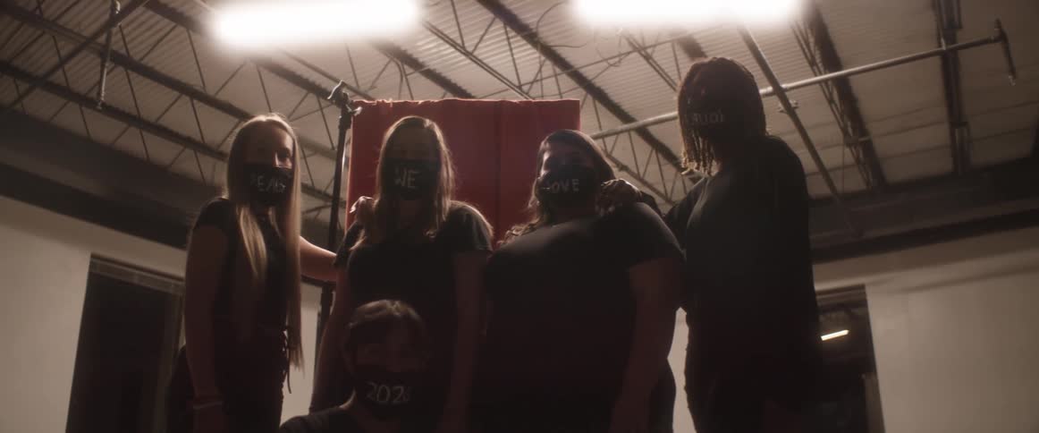 Five women pose in an empty room while wearing black face masks with a written message