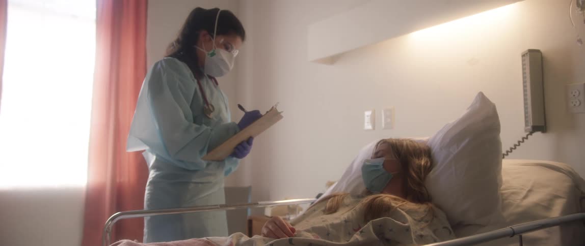 A nurse in personal protective gear approaches the hospital bed of a coughing patient and records the patients condition on a clipboard before exiting the room