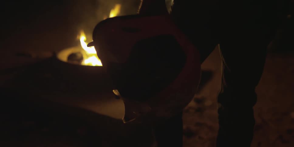 A person holding a white motorbike helmet stands near a fire at night