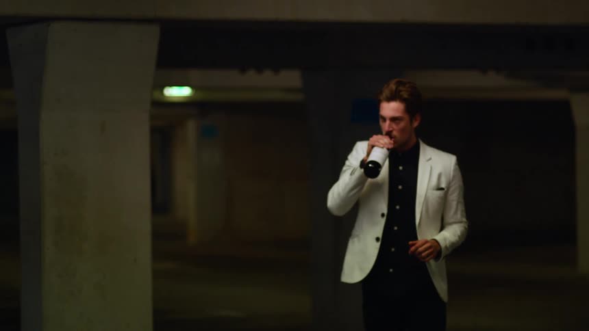 A man walking through a parking garage drinks out of a bottle of wine