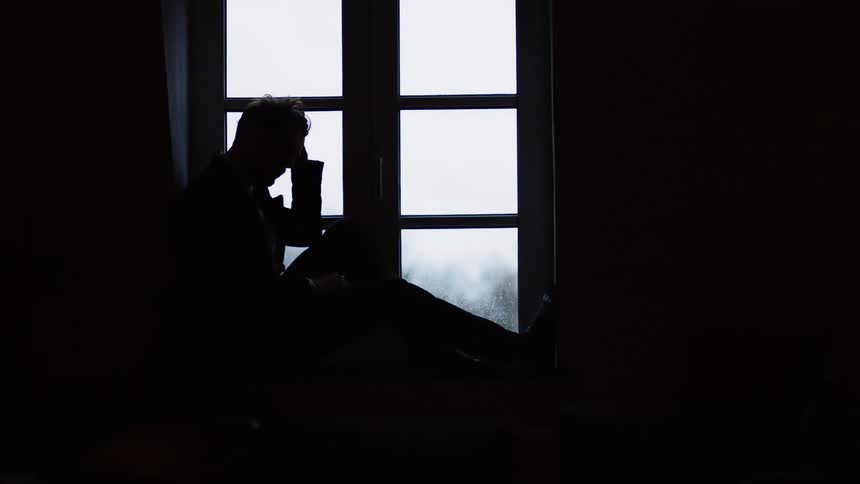 A man sitting in front of a window at home rests his head in his hand and rubs his head