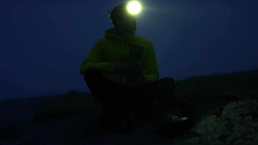 A man with a headlight on sits on a hill and looks at his camera in the dark