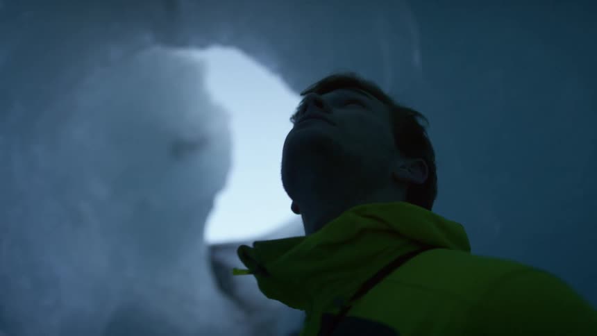 A man standing in a cavern of ice looks up at the sky through a hole in the ceiling