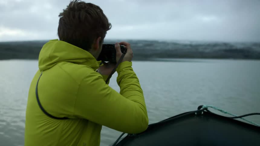A man sits in a raft like boat and holds a camera up to his face to take a photo