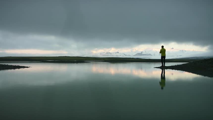 A photographer standing on the edge of a lake looks around him before raising his camera to take a photograph
