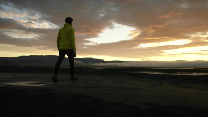 A man with a hoodie and a camera walking near the shore with the sky and clouds in the background