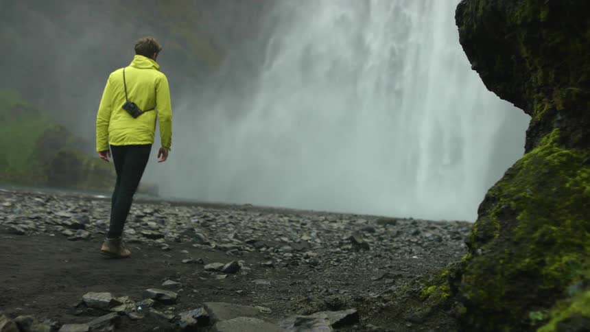 A man with a camera over his shoulder walks towards a waterfall