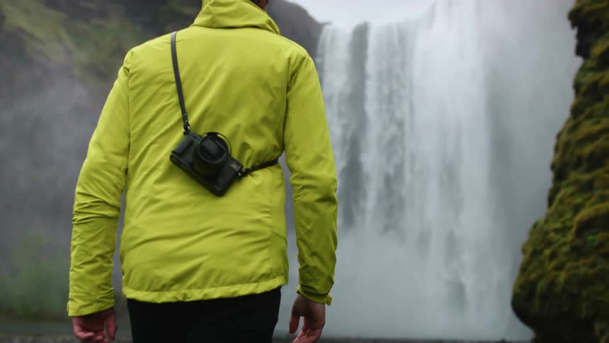 A man with a camera strapped across his shoulder walks to a large waterfall