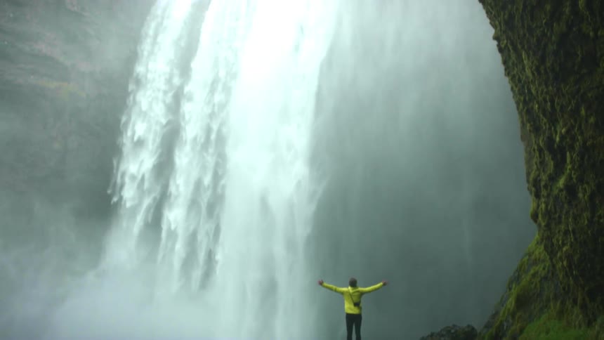 A person stands at the bottom of a large waterfall and puts their arms put beside them