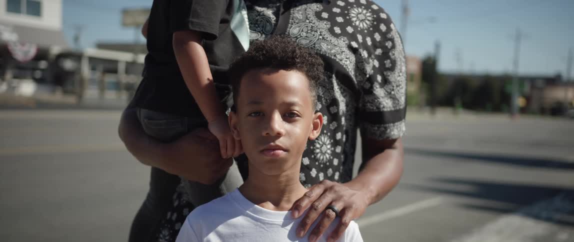 A young boy stands outside with his sibling and his parent while posing