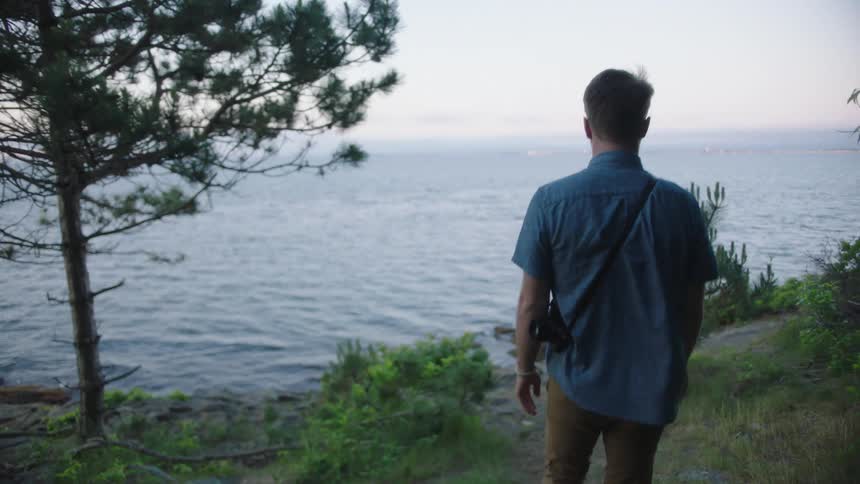 Man carrying his camera over his shoulder walks downhill with the sea in the background