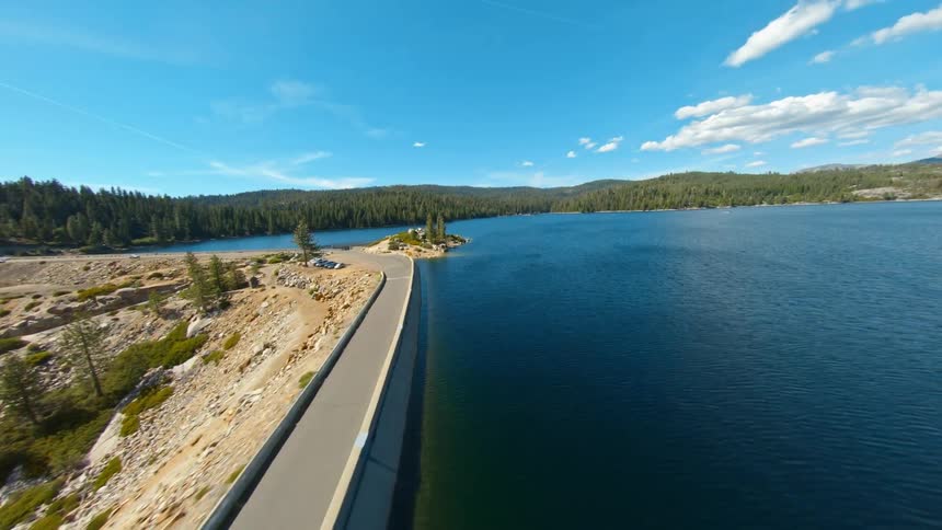 A narrow dam road beside a lake surrounded by a hilly landscape covered in forest