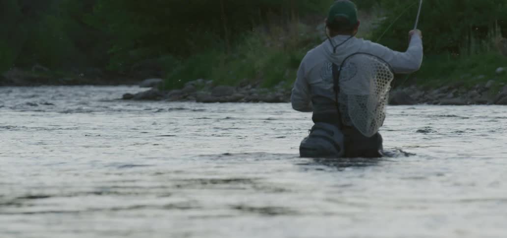 A man with a hat a net and a fishing rod fly fishing in a river