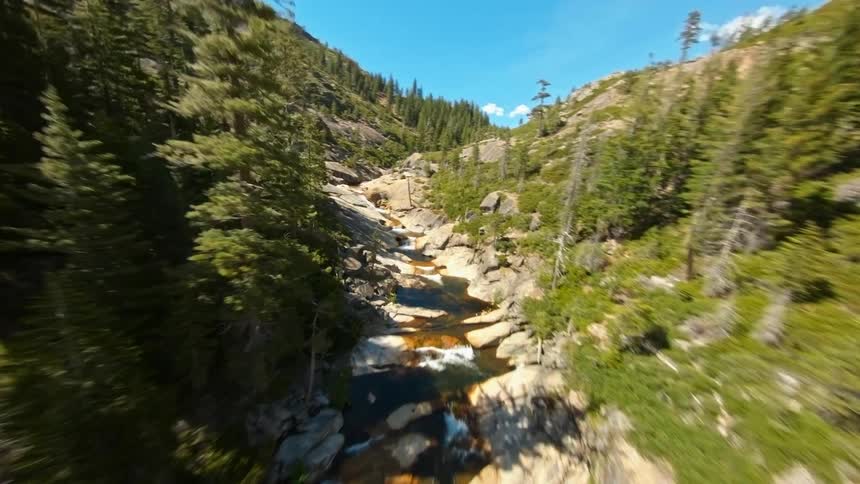 Flying up a stream that runs through the mountains as a forest stands nearby
