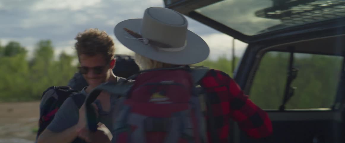 A man and woman retrieve their backpacks from the back of their vehicle and place them on their backs as they prepare to hike through the wilderness