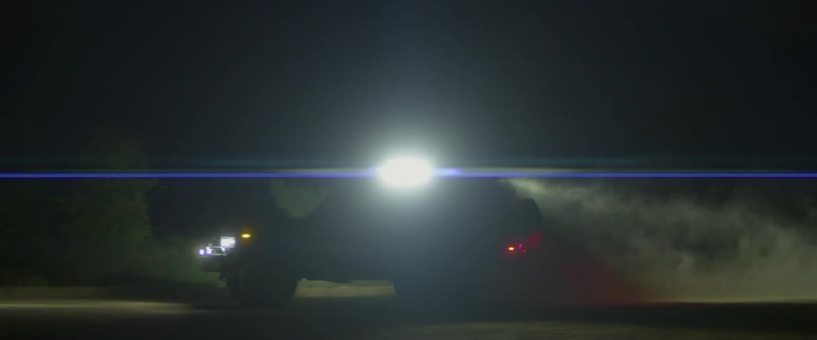 A vehicle with headlights driving by with plants and trees in the background