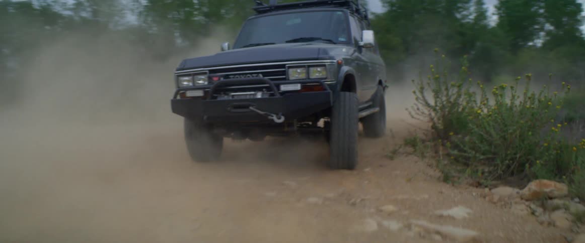 A vehicle creates a cloud of dust behind it as it travels down a dirt road through the countryside on a cloudy day
