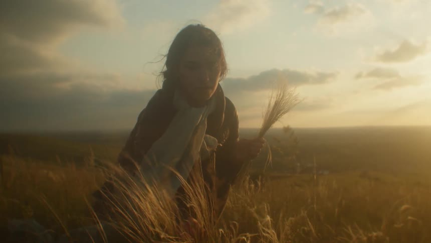 A woman picks and bunches tall grass on a hill as the wind blows and the sun sets