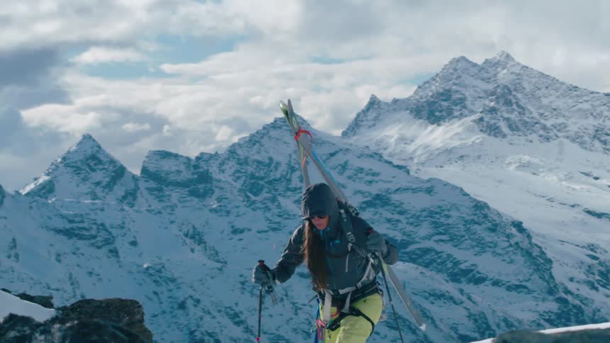 A woman with skis on her back hiking up a mountain with snow with a lake in the background