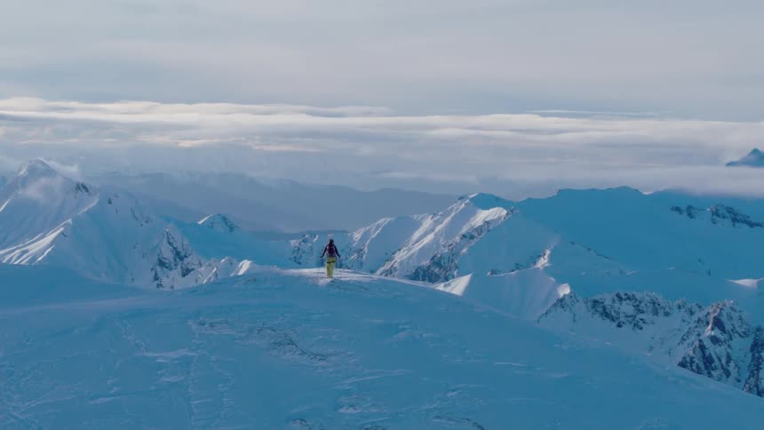 A mountaineer stands atop a mountain and looks out at the snow covered landscape below