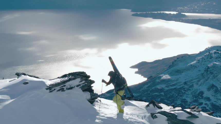 Woman hikes across tall snowy mountain ridge high above lake